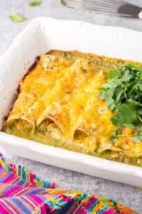 white baking dish filled with green chicken enchiladas topped with orange melted cheese and a mound of bright green cilantro