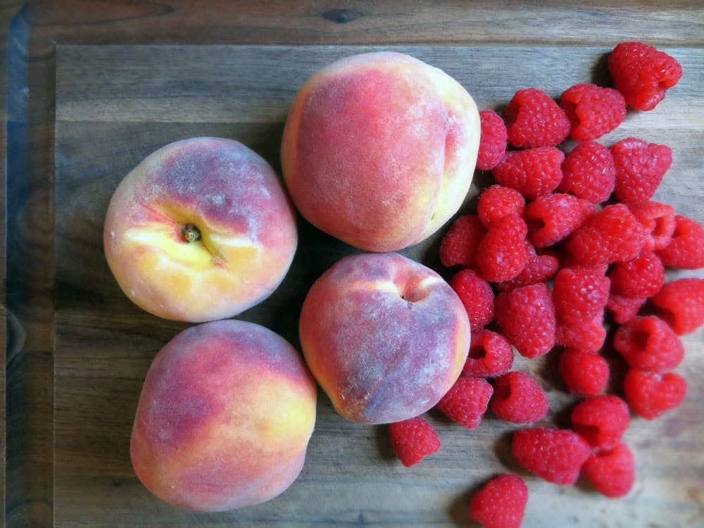 four peaches and a pile of raspberries side by side on a wooden cutting board