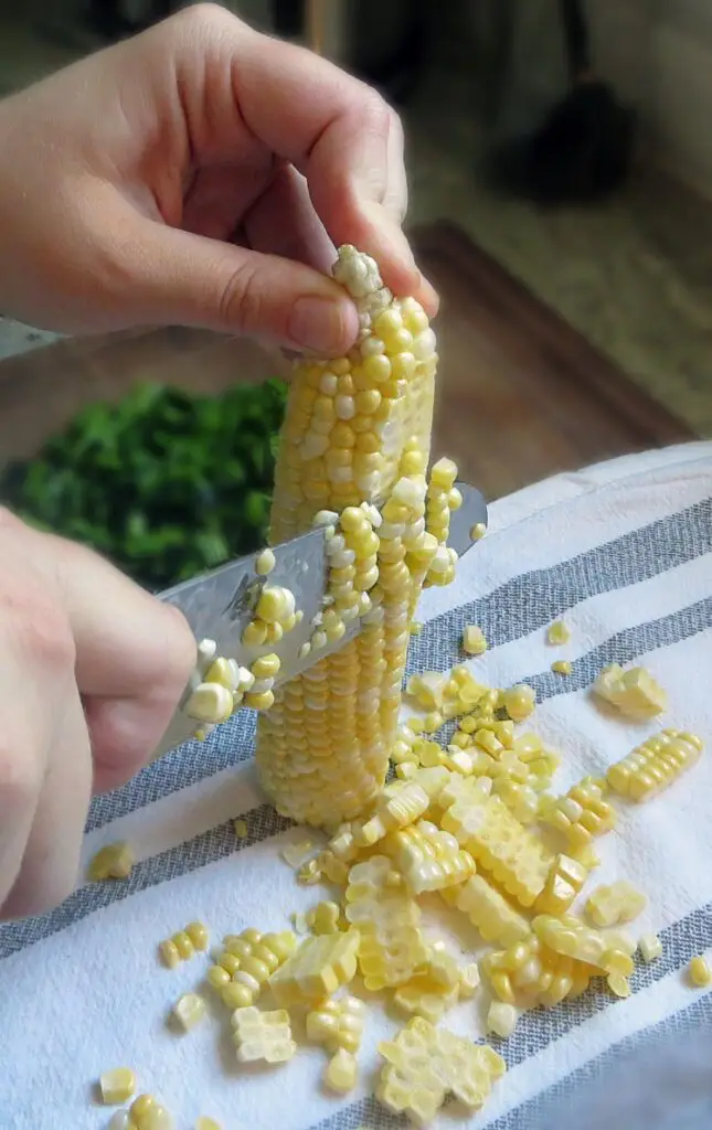 A hand slicing corn kernels from a yellow corn cob with a large chef's knife