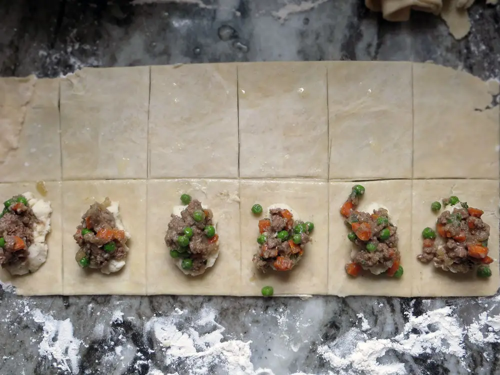 portions of cooked ground meat with vegetables and mashed potatoes on squares of unbaked pastry dough