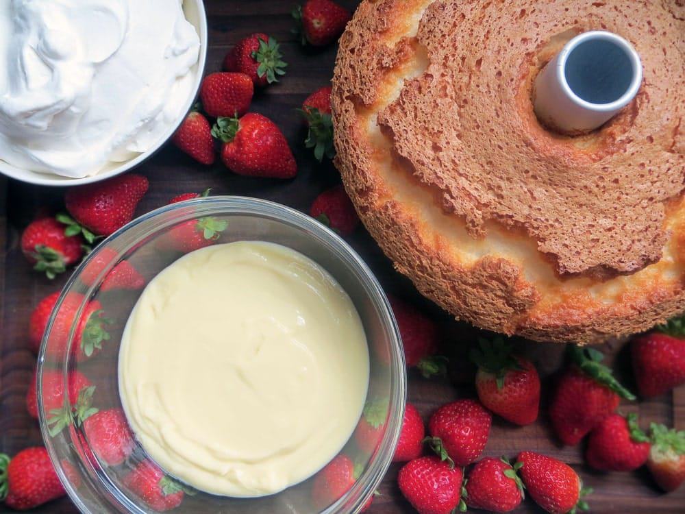 bowls of whipped topping and vanilla pudding beside an angel food cake on a wooden board covered with strawberries