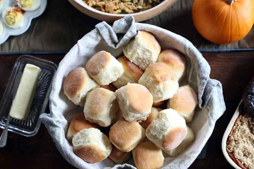 a large linen lined bowl filled with baked yeast rolls surrounded by a Thanksgiving dinner spread