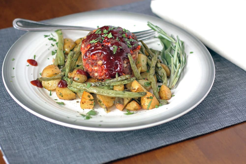 a small barbecued meatloaf on a bed of roasted potatoes and green beans served on a round plate