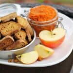 a bowl of baked dog biscuits on a plate with sliced apple and a jar of pumpkin puree