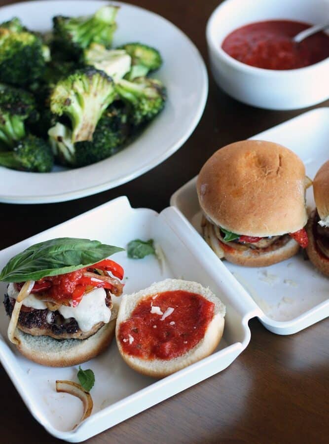 hamburger sliders on buns with marinara sauce, sauteed vegetables, and basil leaves on a white takeout box-style platter