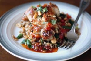 a portion of farro topped with chicken covered in a red sauce and garnished with chopped red vegetables and chopped green herbs