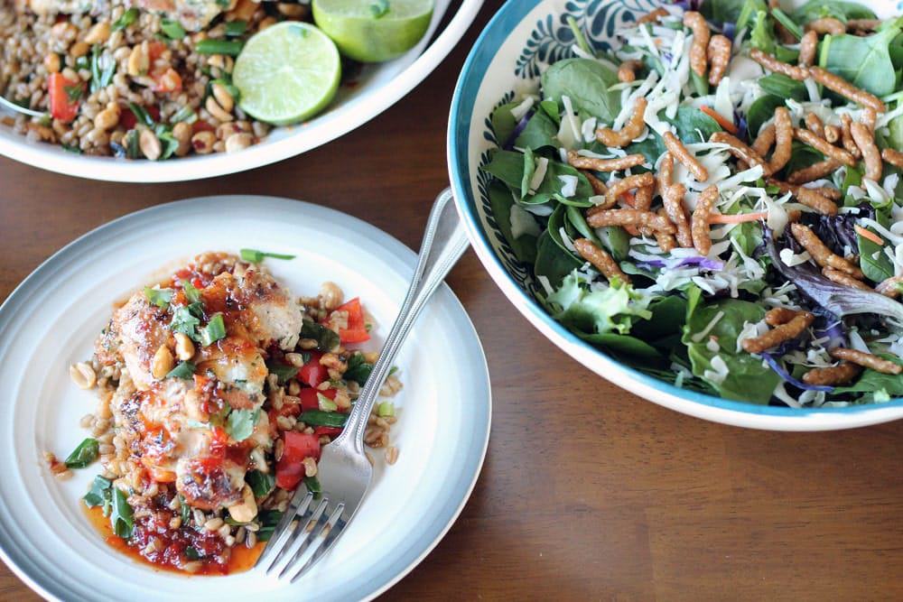 a plate of chicken with vegetables and farro beside a bowl filled with a salad of greens with asian slaw and sesame sticks on a wooden table top