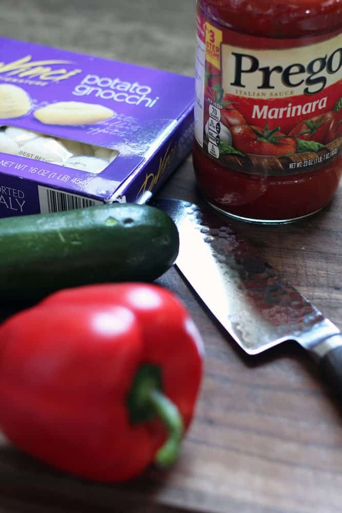 a jar of marinara sauce, a box of potato gnocchi, a zucchini, a red bell pepper, and a sharp looking knife on a wood board