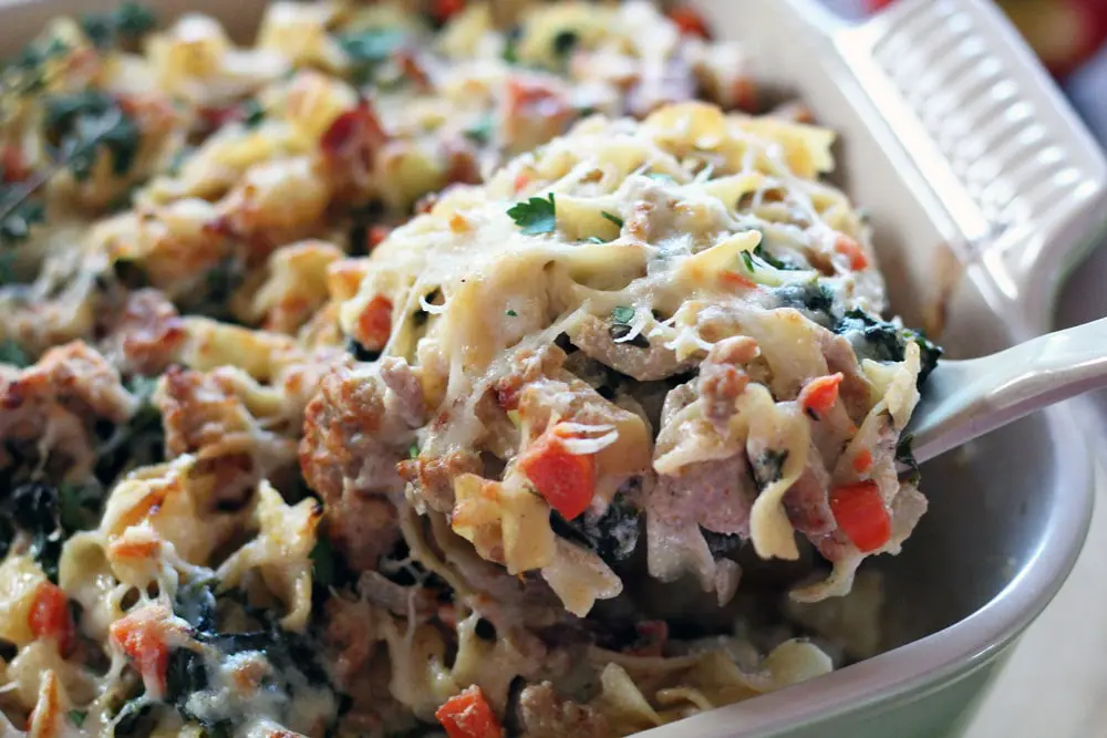 a spoon lifting a portion of a pork, bacon, apple, and pasta casserole from a baking dish