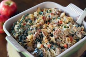 a mixture of pasta with ground meat, bacon, diced apple, and spinach topped with melted cheese in a baking dish