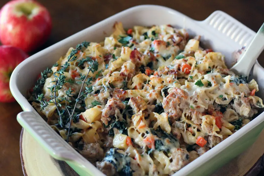 a mixture of pasta with ground meat, bacon, diced apple, and spinach topped with melted cheese in a baking dish