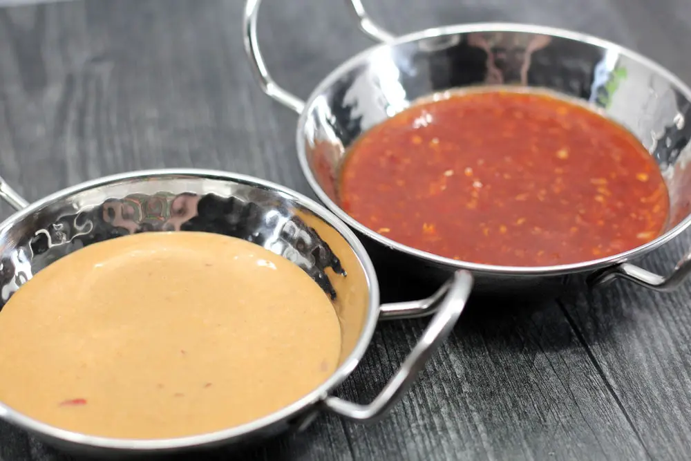 two metal bowls with metal handles; one is filled with a creamy orange sauce, and the other is filled with a red sauce
