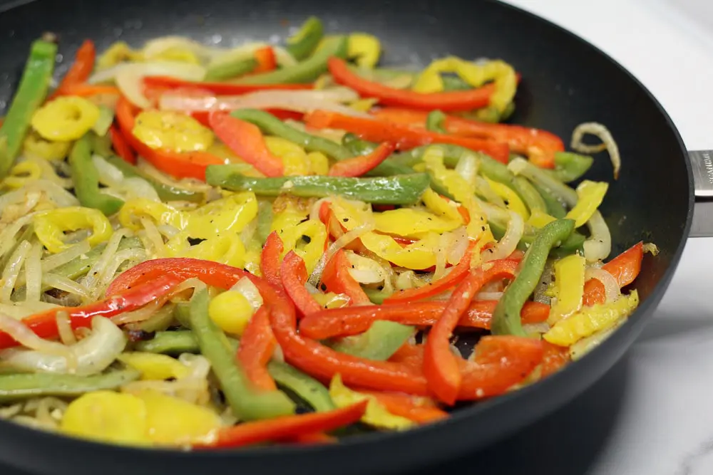 sliced red, green, and yellow peppers with sliced onions sauteing in a black skillet