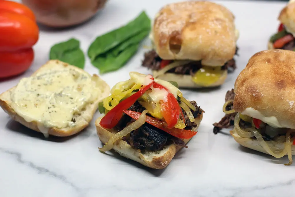 an open bun topped with beef, sauteed peppers and onions, and cheese surrounded by assembled sandwiches with the same ingredients