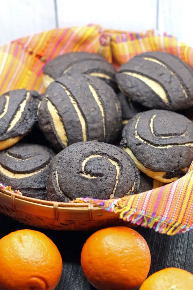 Chocolate Conchas or Mexican Sweet Bread flavored with chocolate and orange in a woven basket lined with a colorful Mexican style linen and surrounded by fresh oranges