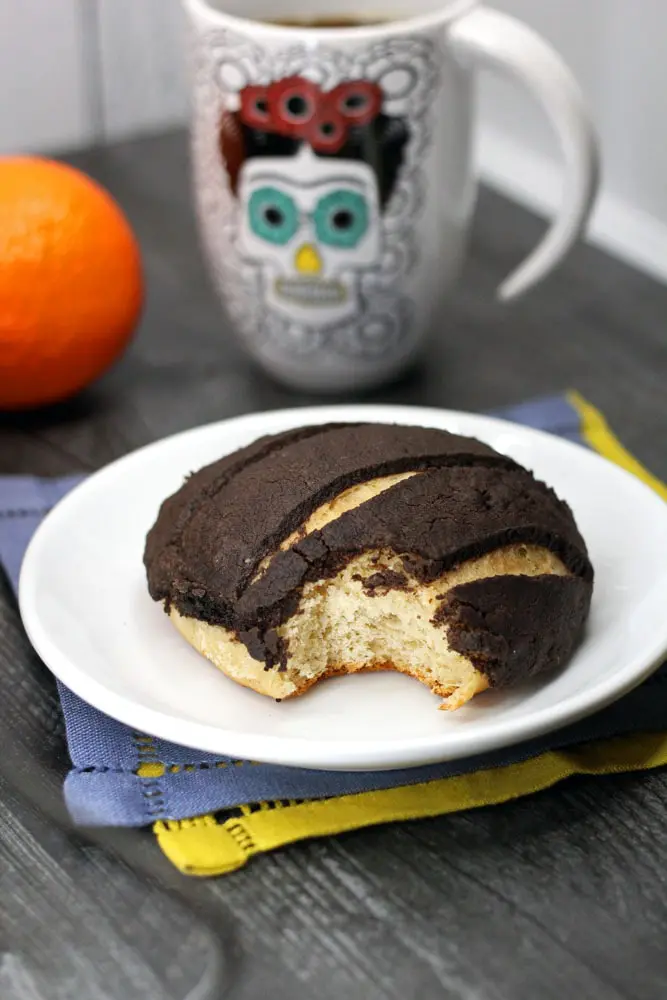 a Chocolate Concha or Mexican Sweet Bread flavored with chocolate and orange on a white plate with a bite taken out of it and a cup of coffee in the background in a day of the dead coffee mug