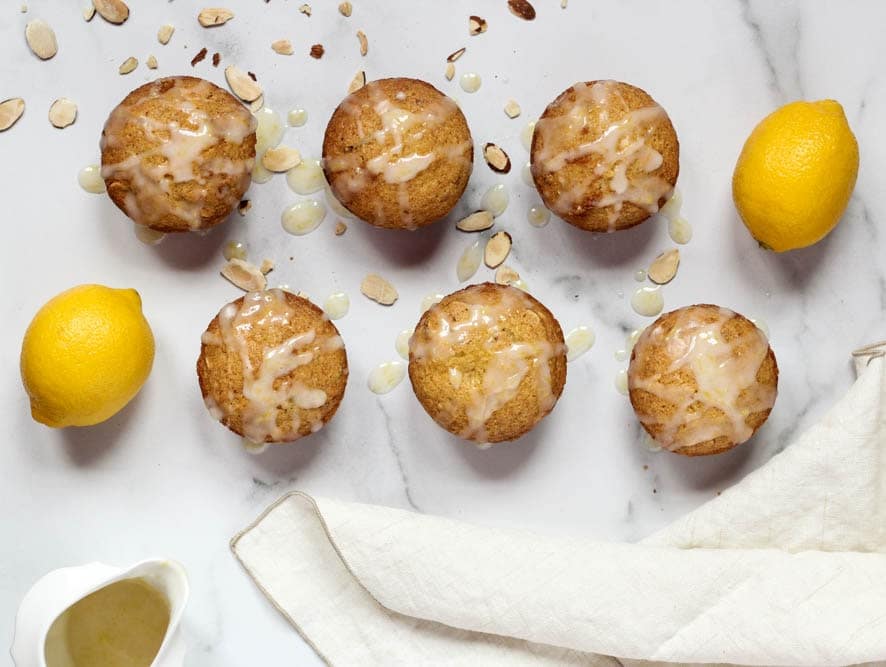 Citrus Almond Muffins evenly spaced on a white marble background with a side of glaze and sprinkled with sliced almonds