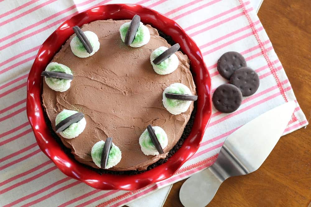 Thin Mint No-Bake Chocolate Cheesecake in a red pie pan garnished with whipped cream, green sprinkles and whole thin mint cookies