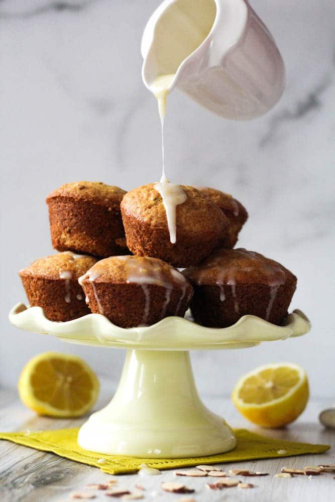 Citrus Almond Muffins on a pale yellow cake pedestal with glaze being poured over the top