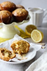 a Citrus Almond Muffin split in half so the inside is visible and a pale yellow cake pedestal with a stack of muffins in the background