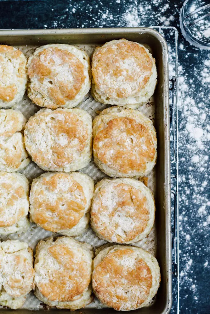 baked Buttermilk Biscuits on a sheet pan sitting on a cooling rack with flour sprinkled across a dark background