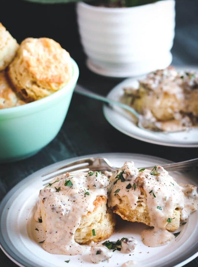 two Buttermilk Biscuits covered in Sausage Gravy with a bowl of biscuits in the background