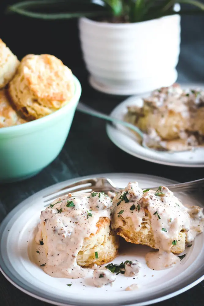 two Buttermilk Biscuits covered in Sausage Gravy with a bowl of biscuits in the background