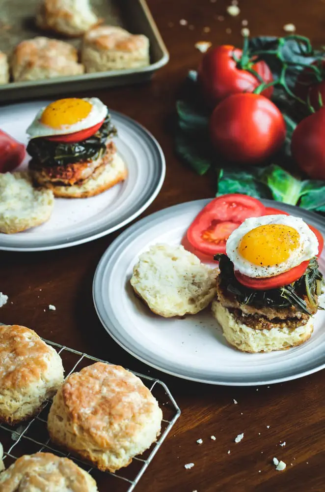 Two Pork and Collard Greens Biscuit Sandwiches served open face to show the sunny side up egg and served with sliced tomato