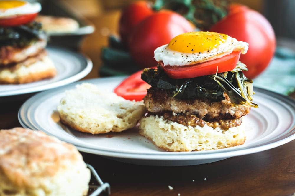 Pork and Collard Greens Biscuit Sandwich with layers of biscuit, whole grain mustard, fried pork chop, cooked collard greens, sliced tomato and a sunny side up egg