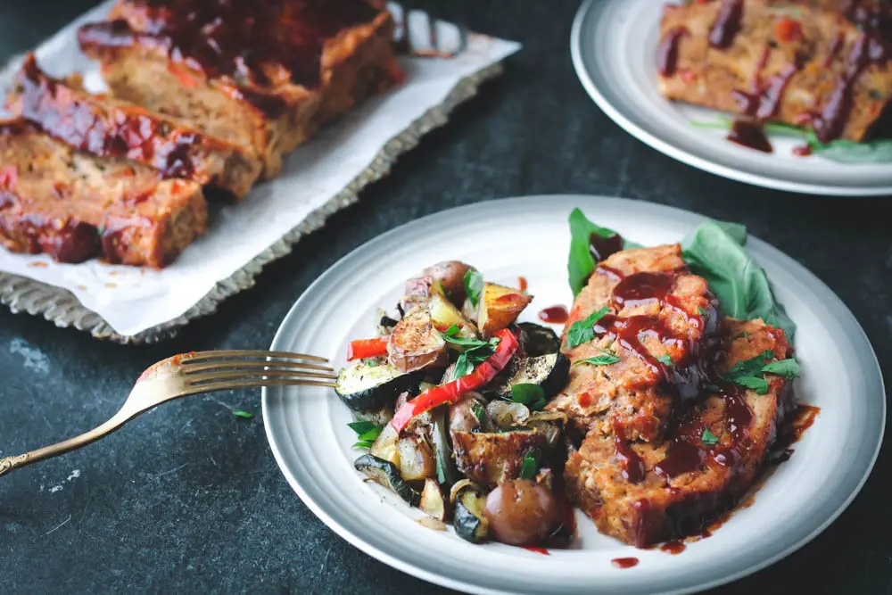 single serving of Sausage and Peppers Meatloaf with Italian Roasted Vegetables served on a white plate with a gray border with the meatloaf in the background