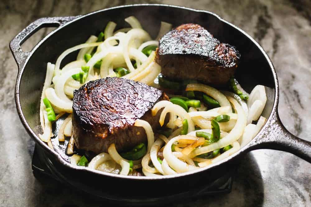 cast iron skillet with two seared steaks and sliced white onions and jalapenos