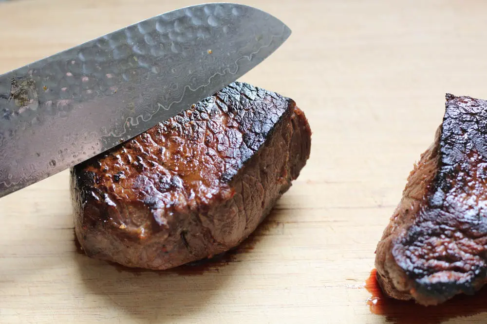closeup shot of a cooked steak on a cutting board with a chef's knife indicating how to cut against the grain of the meat