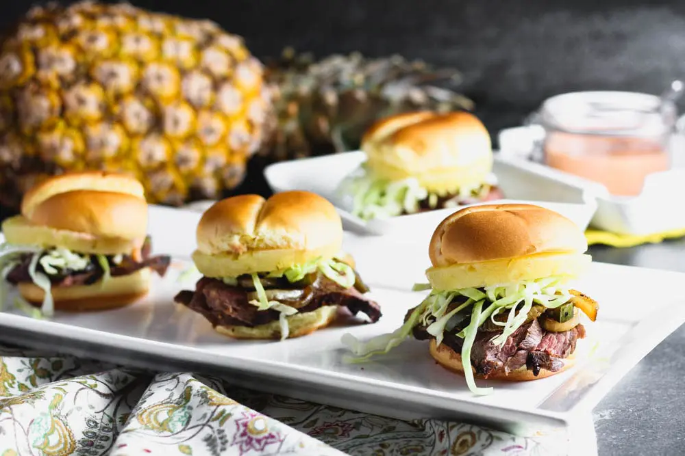 three teriyaki steak and pineapple sliders on a long rectangular white plate with a pineapple and the sweet chili mayo in the background
