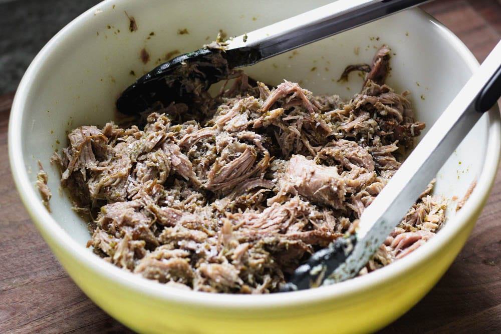 cooked, braised salsa verde pork being shredded with tongs in a large yellow bowl