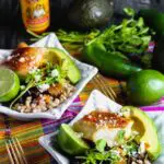 Two bowls of cooked farro topped with taco seasoned cod, fresh avocado, lime, black beans, cilantro and cooked vegetables with hot sauce and lime in the background