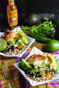 Two bowls of cooked farro topped with taco seasoned cod, fresh avocado, lime, black beans, cilantro and cooked vegetables with hot sauce and lime in the background