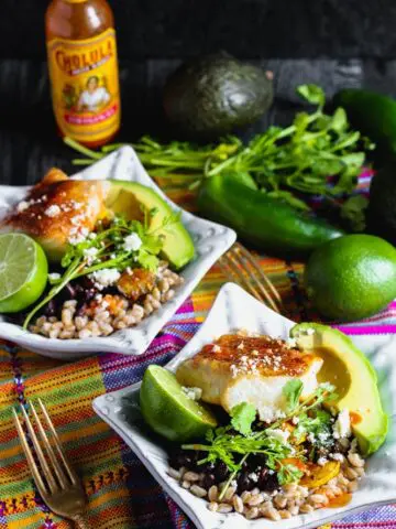 Two bowls of cooked farro topped with taco seasoned cod, fresh avocado, lime, black beans, cilantro and cooked vegetables with hot sauce and lime in the background