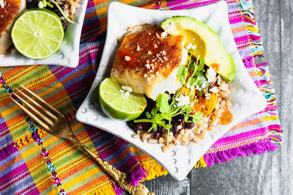 closeup of a bowl of cooked farro topped with taco seasoned cod, fresh avocado, lime, black beans, cilantro and cooked vegetables