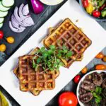 overhead shot of falafel waffles topped with fresh mint and surrounded by red onions, tomatoes and lamb sausage