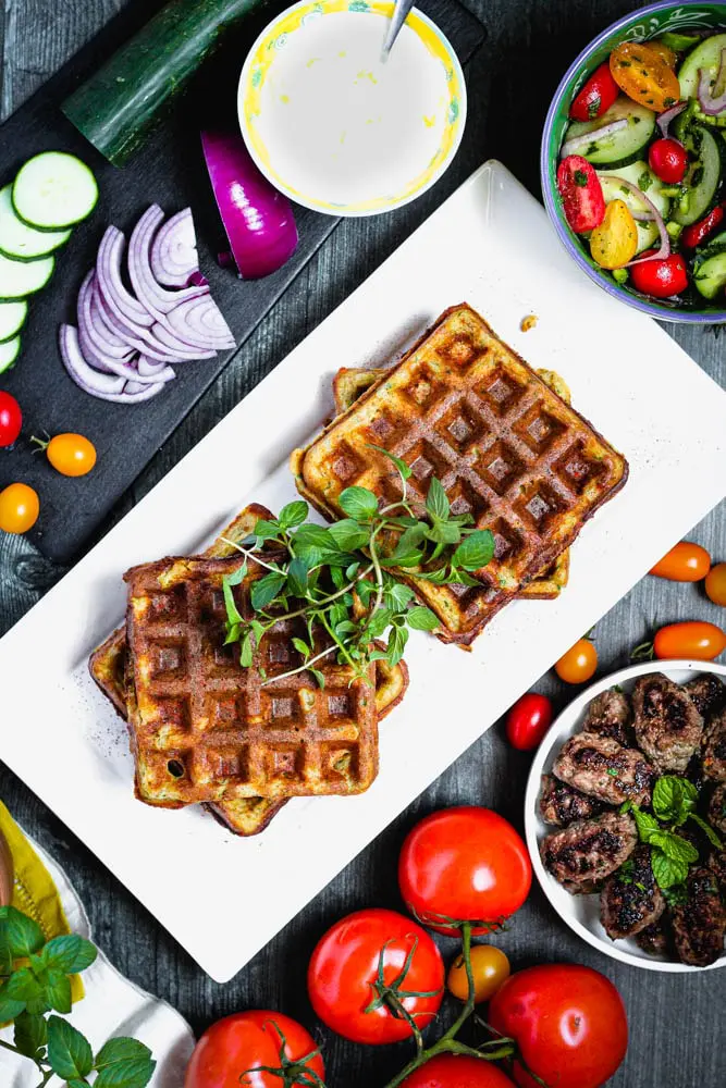 overhead shot of falafel waffles topped with fresh mint and surrounded by red onions, tomatoes and lamb sausage