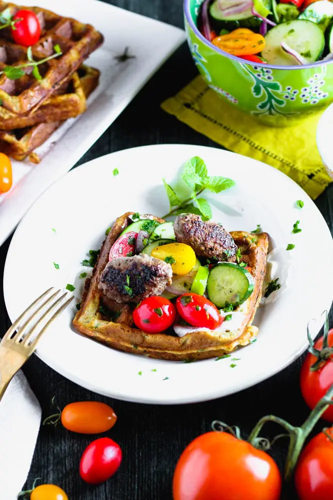 dressed falafel waffle that is topped with yogurt sauce, cucumber tomato salad and lamb sausage