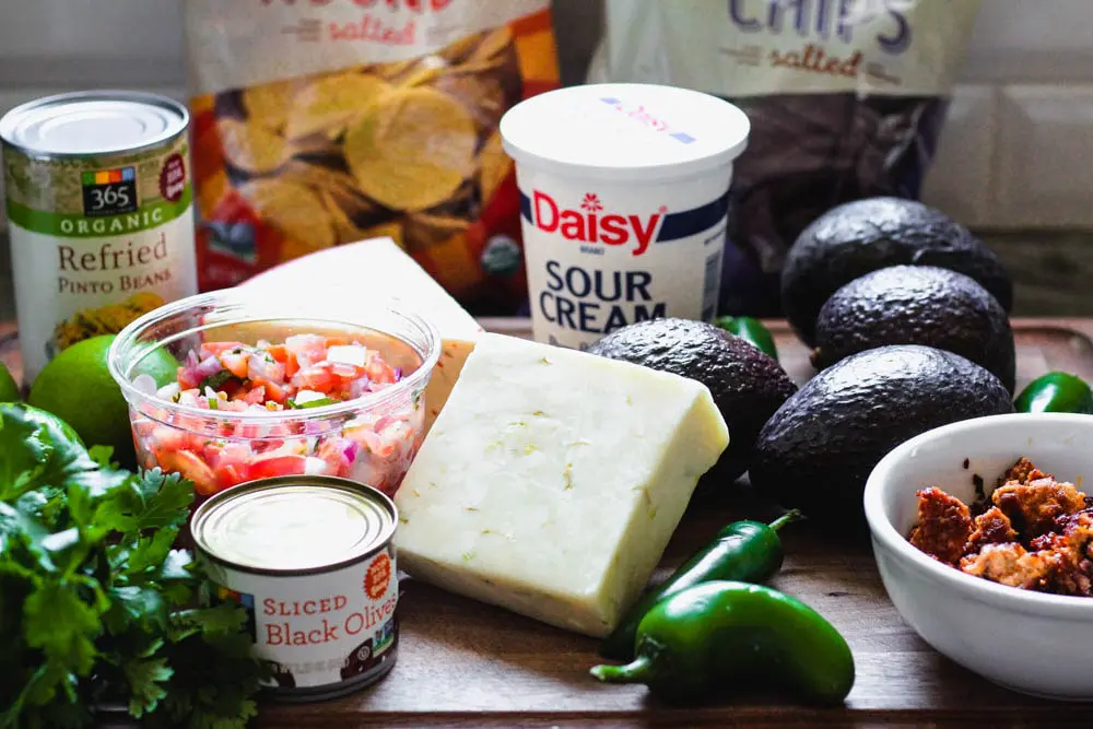 ingredients for seven layer dip including pico de gallo, canned refried beans and sliced olives, sour cream, avocado, block of pepper jack, jalapenos and cooked chorizo