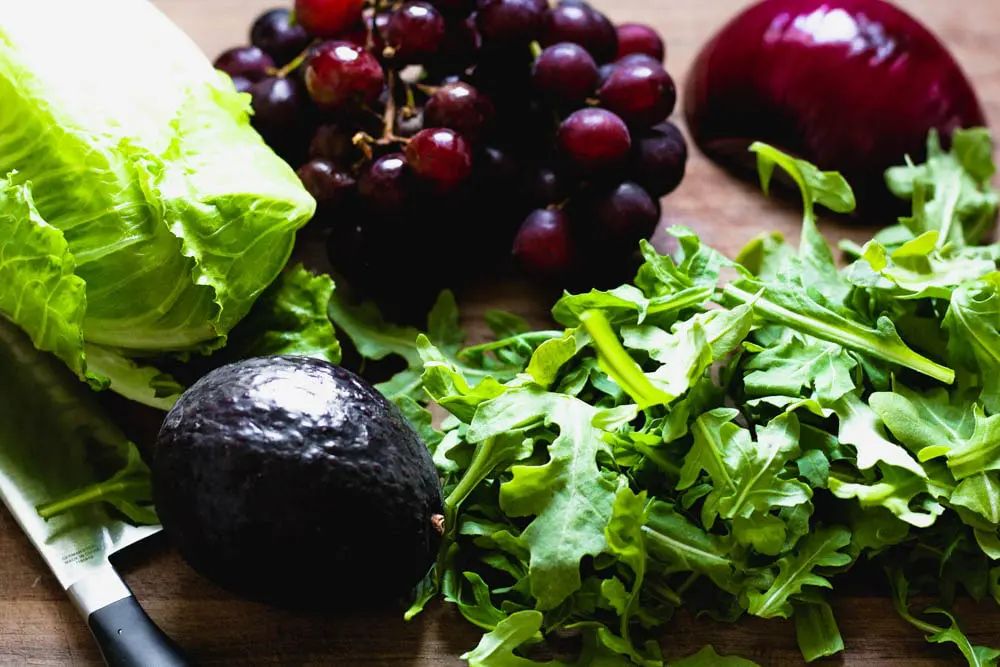 Raw salad ingredients before preparing including romaine lettuce, grapes, a whole avocado, arugula and half a red onion