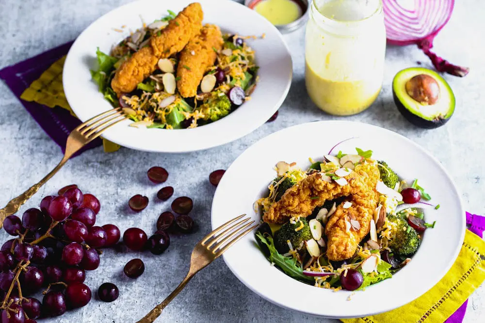 Two bowls of salad topped with roasted broccoli, sliced grapes and almonds and shredded cheese with a side of honey mustard