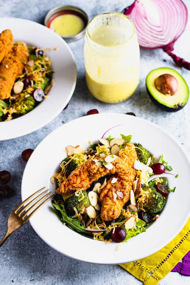 Bowl of salad topped with roasted broccoli, sliced grapes and almonds and shredded cheese with a side of honey mustard