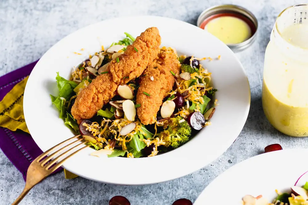 Bowl of salad topped with roasted broccoli, sliced grapes and almonds and shredded cheese with a side of honey mustard