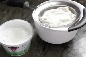 greek yogurt sitting next to the greek yogurt straining in a fine strainer over a small mixing bowl to catch the extra liquid