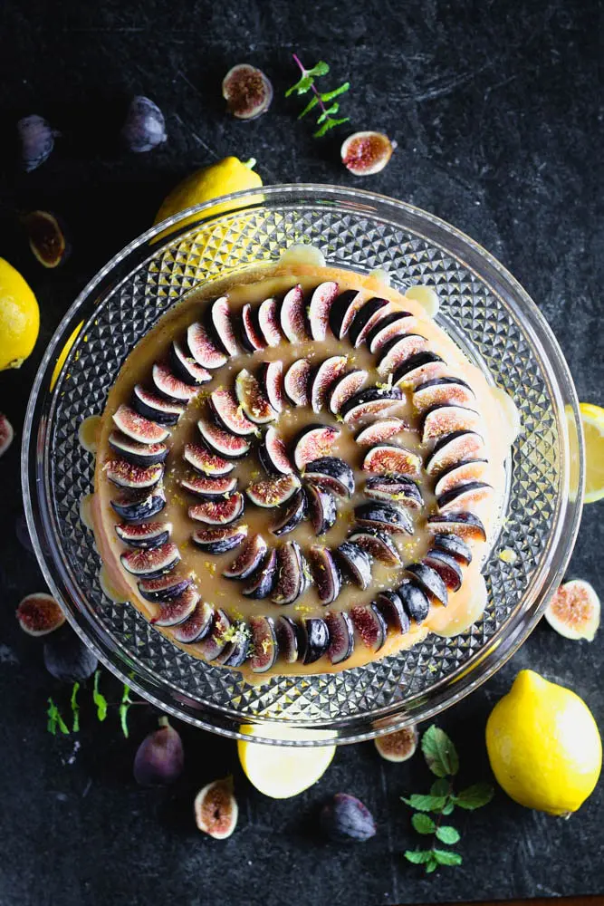 overhead shot of lemon yogurt cake topped with honey caramel and quartered figs on a crystal cake pedestal with lemons and figs in the background
