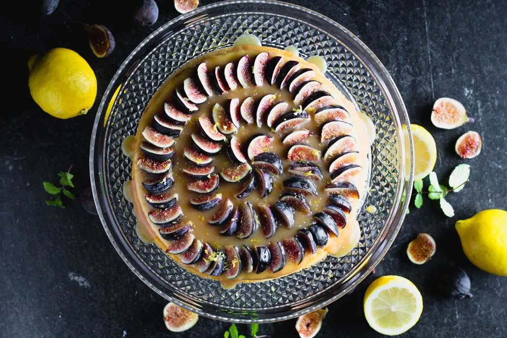 overhead shot of lemon yogurt cake topped with honey caramel and quartered figs on a crystal cake pedestal with lemons and figs in the background