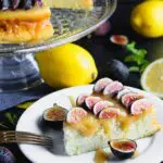 Closeup photo of a slice of lemon yogurt cake topped with honey caramel and quartered figs on a white plate with a crystal cake pedestal with the yogurt cake in the background along with lemons and figs